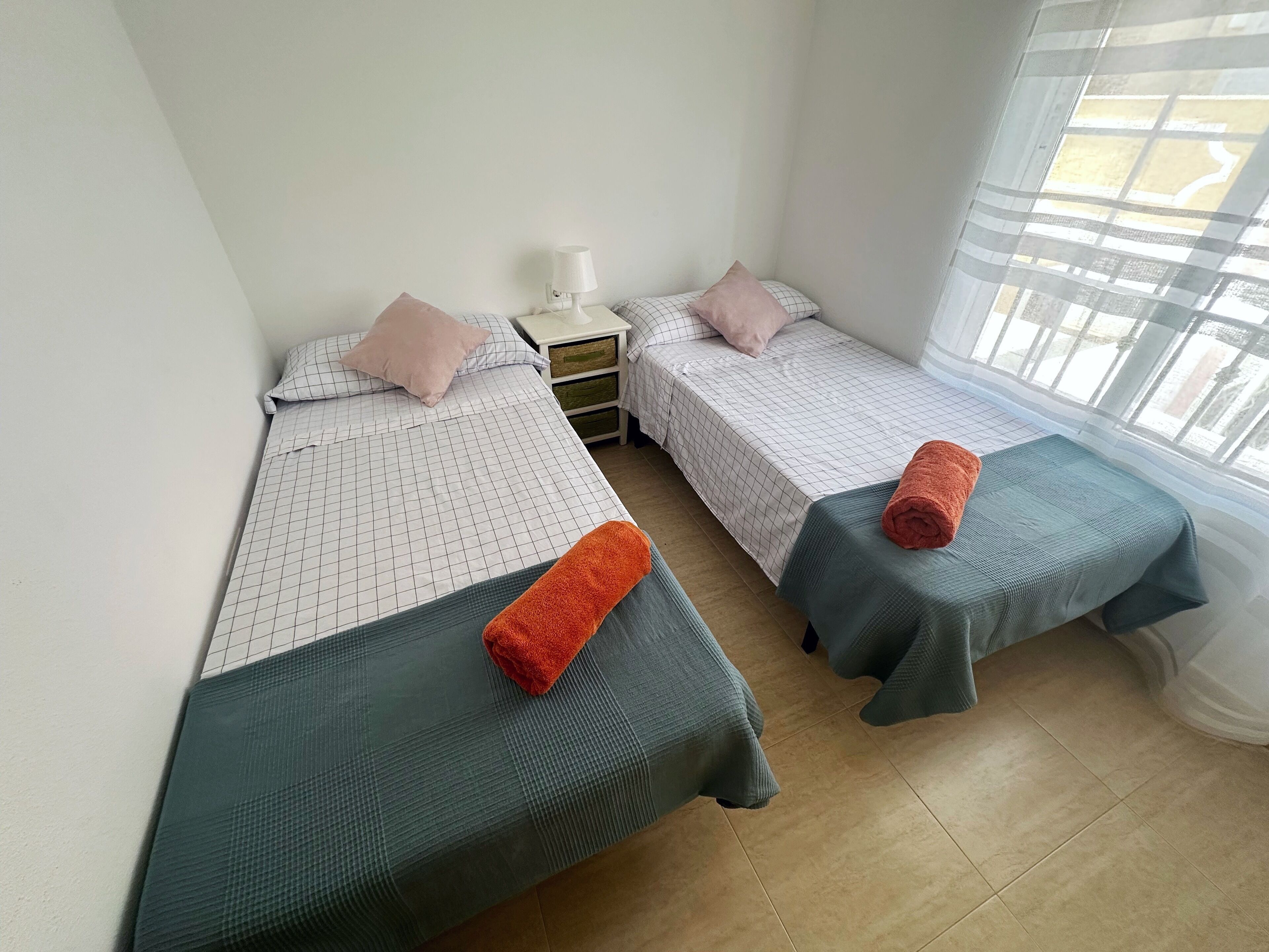 A room with two beds, each with a pillow and a rolled towel. There is a nightstand with a lamp between the beds.