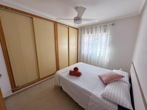 Egy hálószoba, amelyben van egy ágy, egy mennyezeti ventilátor, egy függönnyel ellátott ablak és egy szekrény.