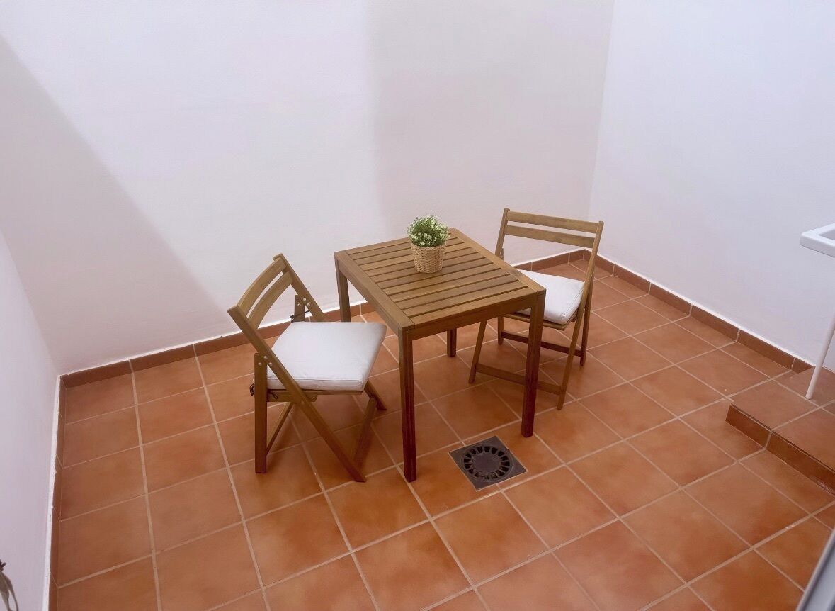 Una habitación pequeña con paredes de azulejos, una mesa de madera y dos sillas, una planta en maceta sobre la mesa, y una pared blanca.