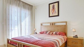 Quarto com cama de madeira, mesa de cabeceira com abajur e uma grande janela com cortinas finas.