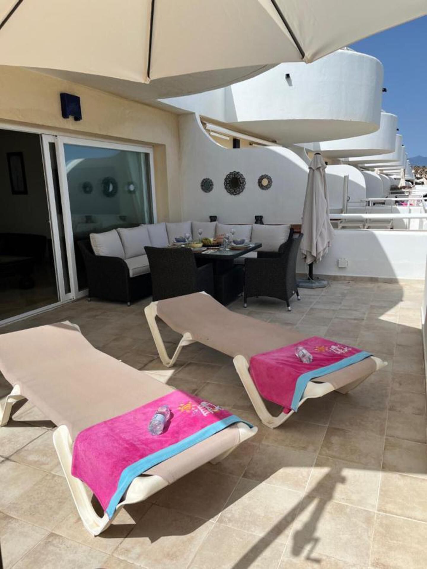 A sunlit outdoor seating area with lounge chairs and a table, a sliding glass door, and decorative wall elements.