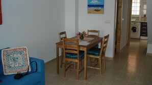 Un coin repas avec une table et des chaises en bois, un canapé bleu agrémenté d’un coussin décoratif et, au mur, un tableau représentant une scène de plage.