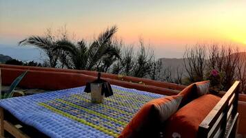 A bed with a blue quilt and a black object on it, set against a sunset view with palm trees and hills in the background.