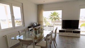 A modern living room with a glass dining table, white chairs, a piano, a TV, and a view of palm trees. - 3-bed Villa Sleeps 8 With Private Pool (Santa Pola)