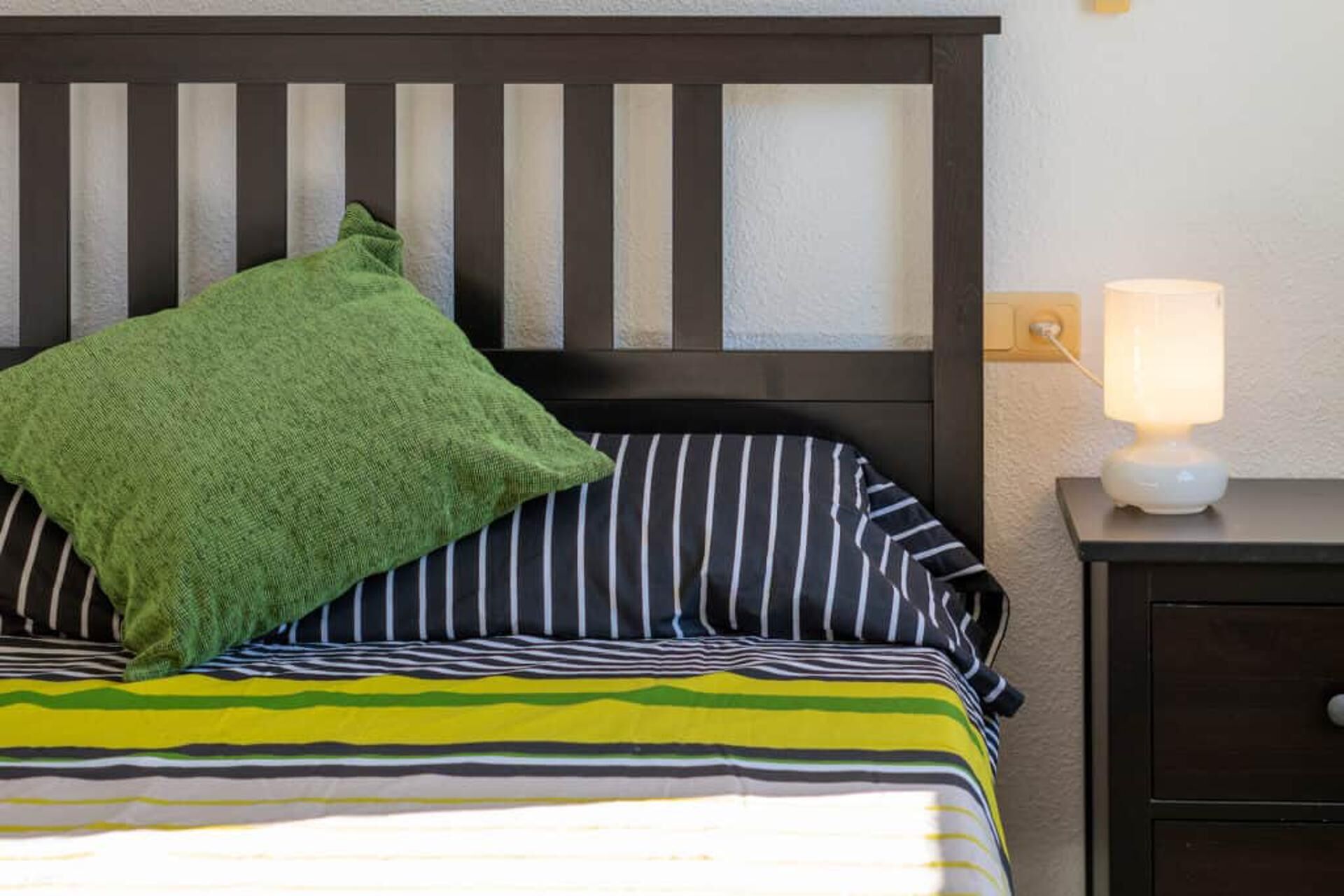 A bed with striped bedding, a green pillow, and a bedside lamp.