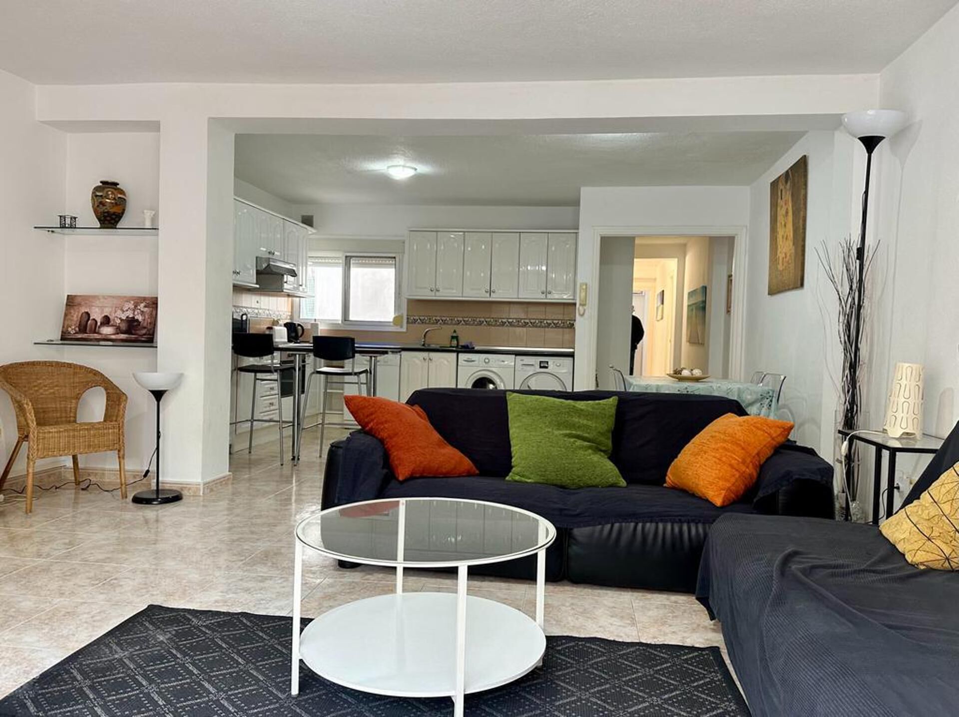 Una sala de estar moderna con un sofá negro, una mesa de centro de vidrio y un comedor con armarios blancos y una cocina al fondo.
