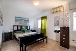 Un dormitorio con cama, cómoda, mesita de noche, un cuadro, ventana con cortinas y equipo de aire acondicionado.