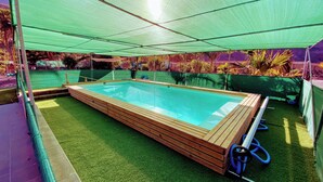 Une piscine rectangulaire dotée d’une terrasse en bois, entourée de gazon synthétique et surplombée par une tonnelle verdoyante.