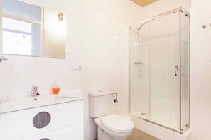 Egy modern fürőszoba, üvegfalas zuhanyzóval, fehér mosdókagylóval, mely felett tükör található, és fehér fedéllel ellátott WC-vel.