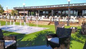 Un espacio al aire libre con varias mesas y sillas, una fuente de agua y un edificio con amplios ventanales al fondo.