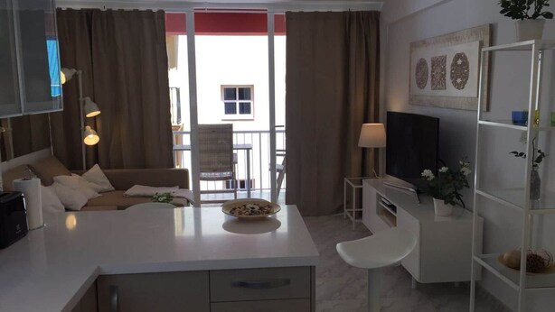 A modern living room with a sofa, a dining table, and a TV. There is a balcony with a chair and a plant. - Apartment Sleeps 4 Near Beach (Fuengirola)