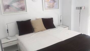 Una cama bien tendida con ropa de cama blanca y tres almohadas, dos negras y una beige, en una habitación con paredes blancas y una lámpara de pie.