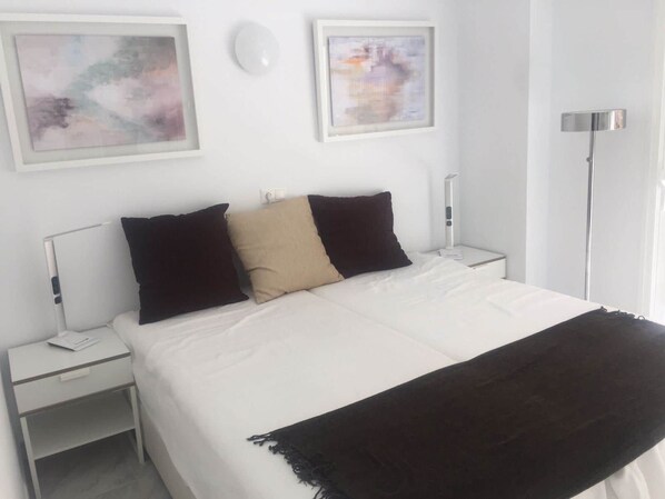 A neatly made bed with white bedding and three pillows, two black and one beige, in a room with white walls and a floor lamp. - Apartment Sleeps 4 Near Beach (Fuengirola)