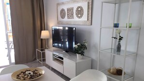 A modern living room with a flat-screen TV on a white cabinet, a dining table with a bowl of snacks, a white chair, and a glass shelf with decorative items. - Apartment Sleeps 4 Near Beach (Fuengirola)