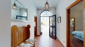 A narrow hallway with a wooden bench, a bed with a blue bedspread, a large mirror, and a view of the outside through a window and door.