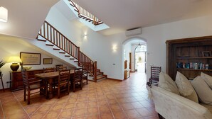 A spacious living area with a wooden staircase, a dining table with chairs, a sofa, and a bookshelf.