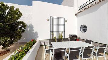 Un patio con una mesa y sillas blancas, una parrilla y un pequeño jardín con plantas y flores.