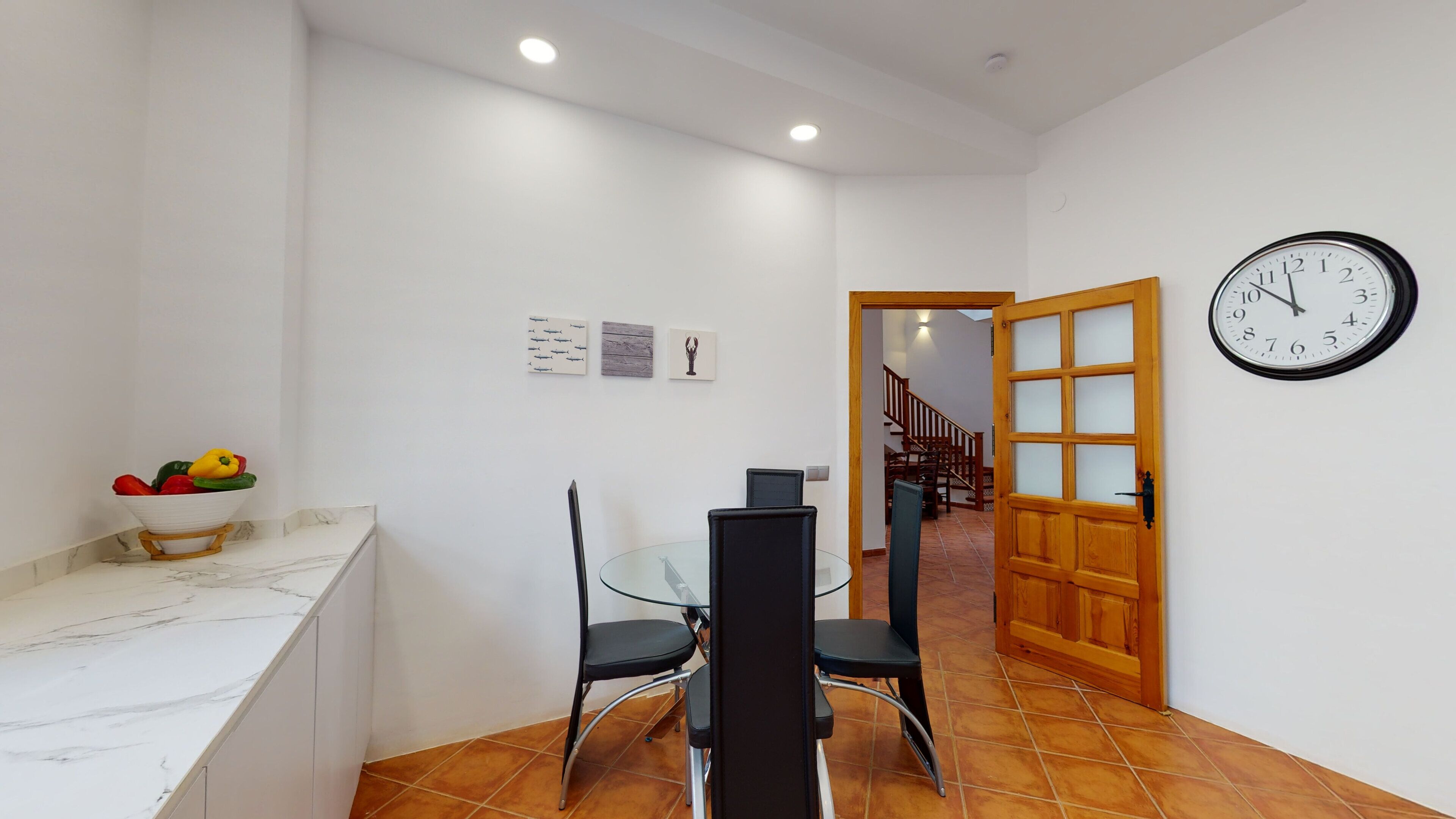A kitchen with a marble countertop, a bowl of fruit, a dining table with chairs, and a clock on the wall.