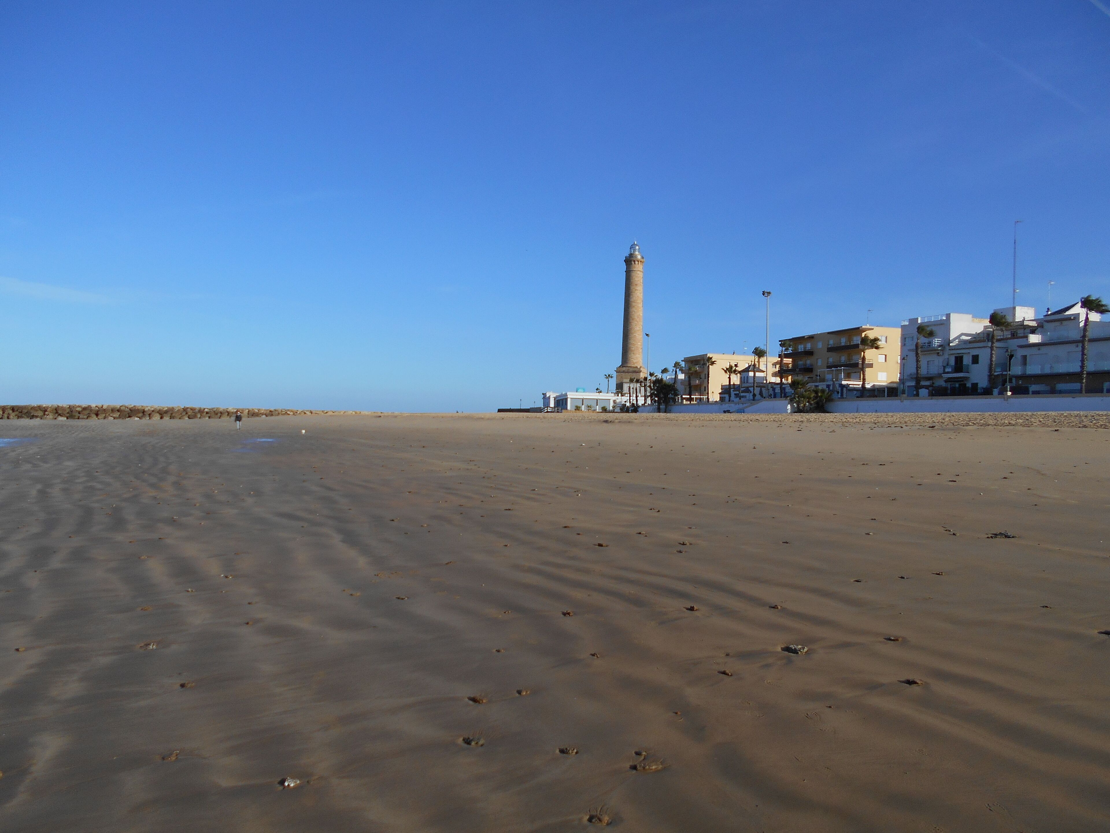 A sandy beach with a tall tower, buildings, and palm trees.
