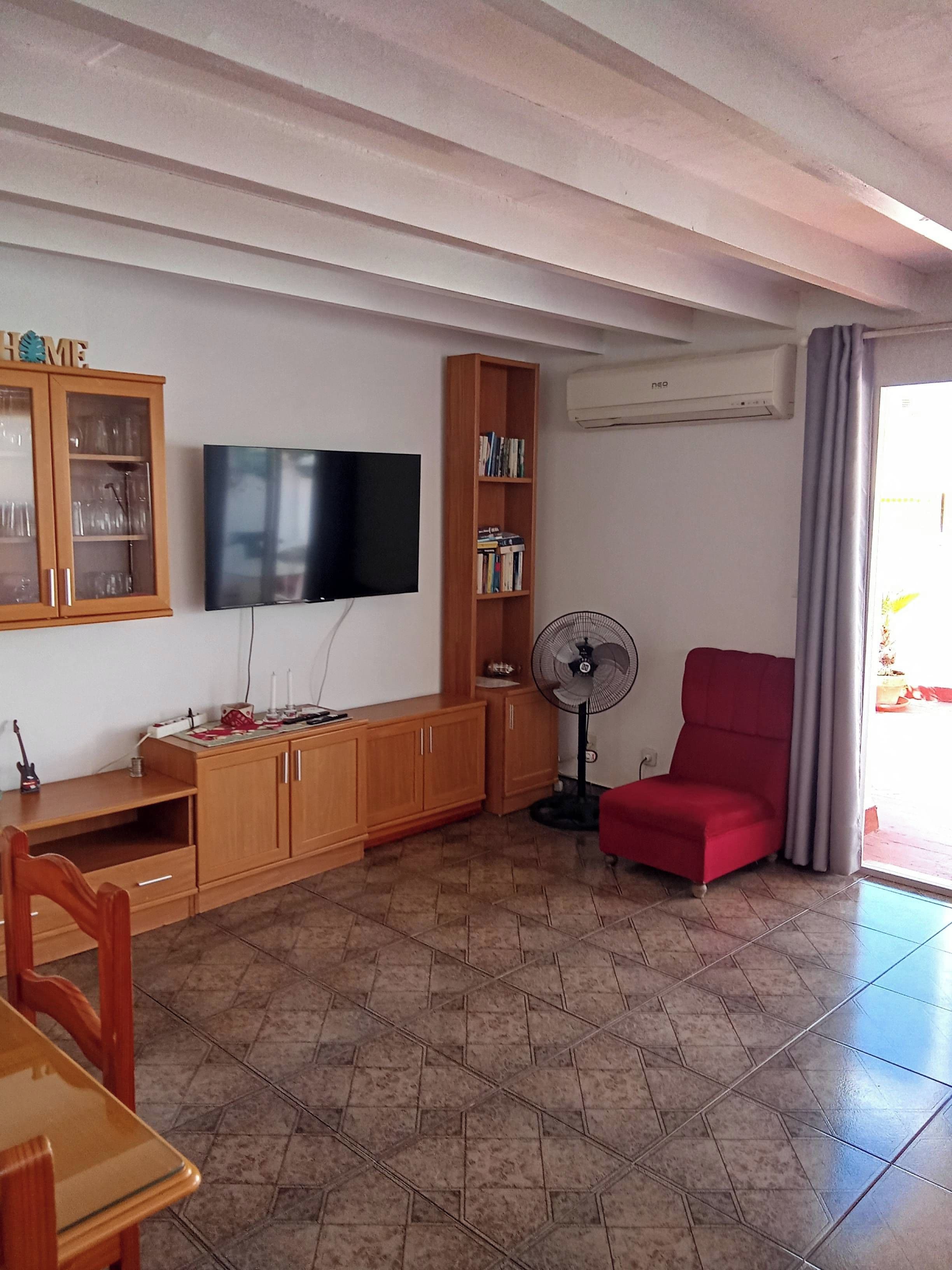 Un soggiorno con televisione, mobiletto in legno, libreria, poltrona rossa e una ventola.