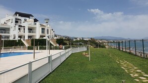 A modern white building with a swimming pool, a fenced grassy area, and a view of the sea. - Captivating 2-bed Apartment First Line Beach (Casares)