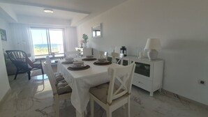 A dining area with a table set for four, white chairs, a white cabinet with a lamp, and a view of the outdoors through a large window. - Captivating 2-bed Apartment First Line Beach (Casares)