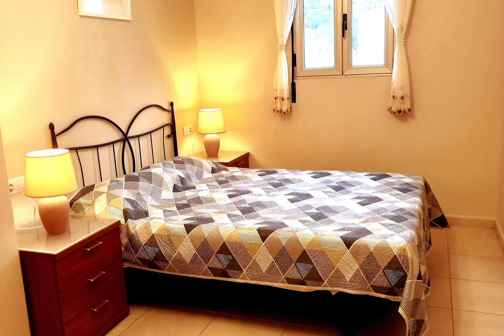 Un dormitorio con cama, dos lámparas de noche, una mesita de noche de madera y una ventana con cortinas.