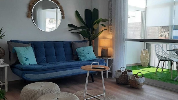 A modern living room with a blue sofa, round ottomans, a small wooden table, and a large window offering a view of a green outdoor area. - Apartment Fuengirola Carvajal Beachfront Malaga (Fuengirola)