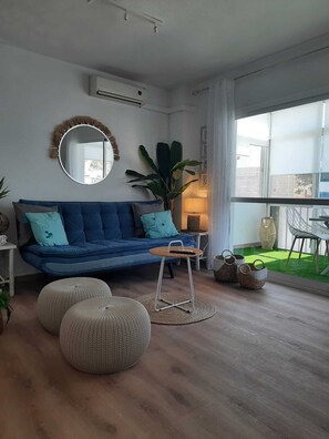 Un salon moderne avec un canapé bleu, des poufs ronds, une petite table en bois et une grande fenêtre donnant sur un espace extérieur verdoyant.