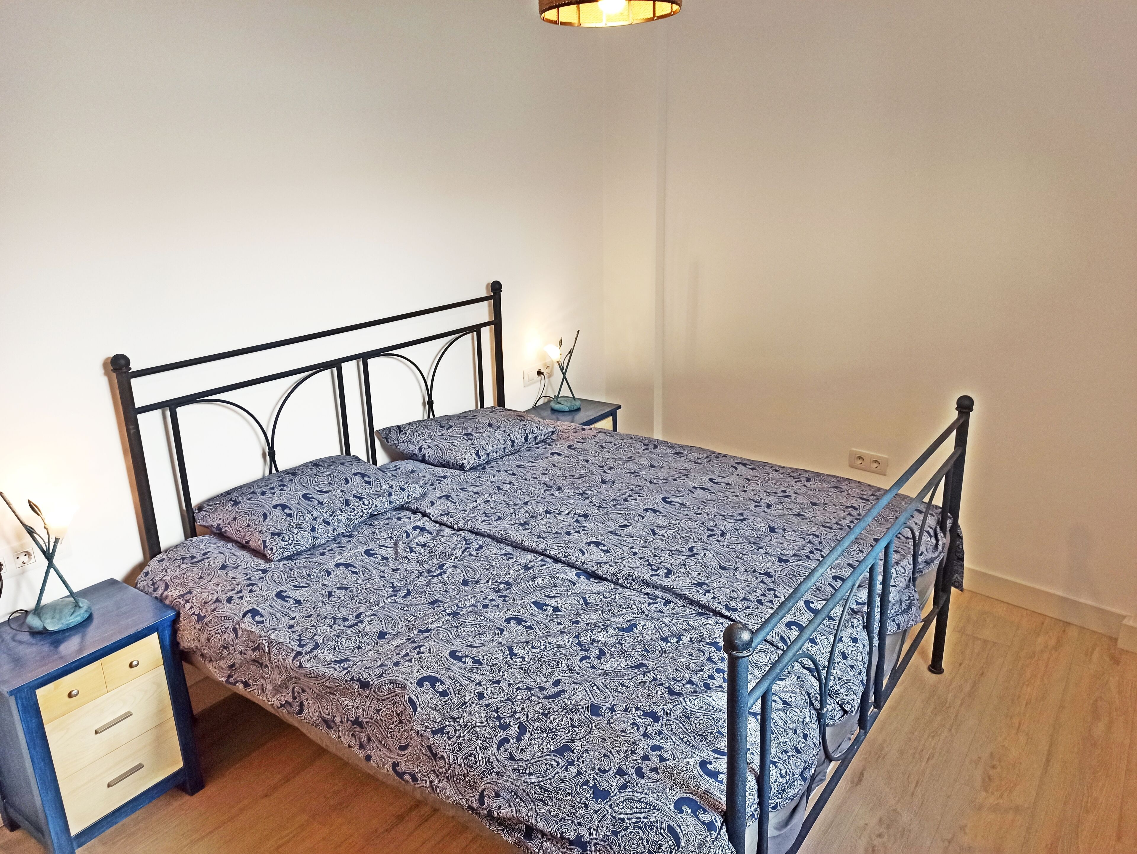 Un lit simple avec un cadre en métal et une couvre-lit à motifs, une table de chevet avec des tiroirs et une prise électrique murale.
