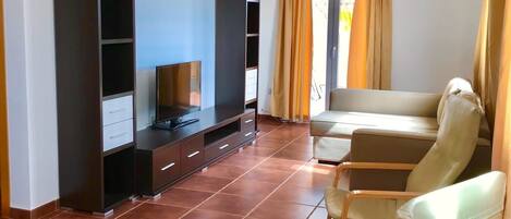A spacious living room with a flat-screen TV, a sofa, and two armchairs. There are built-in shelves and a large window with curtains.