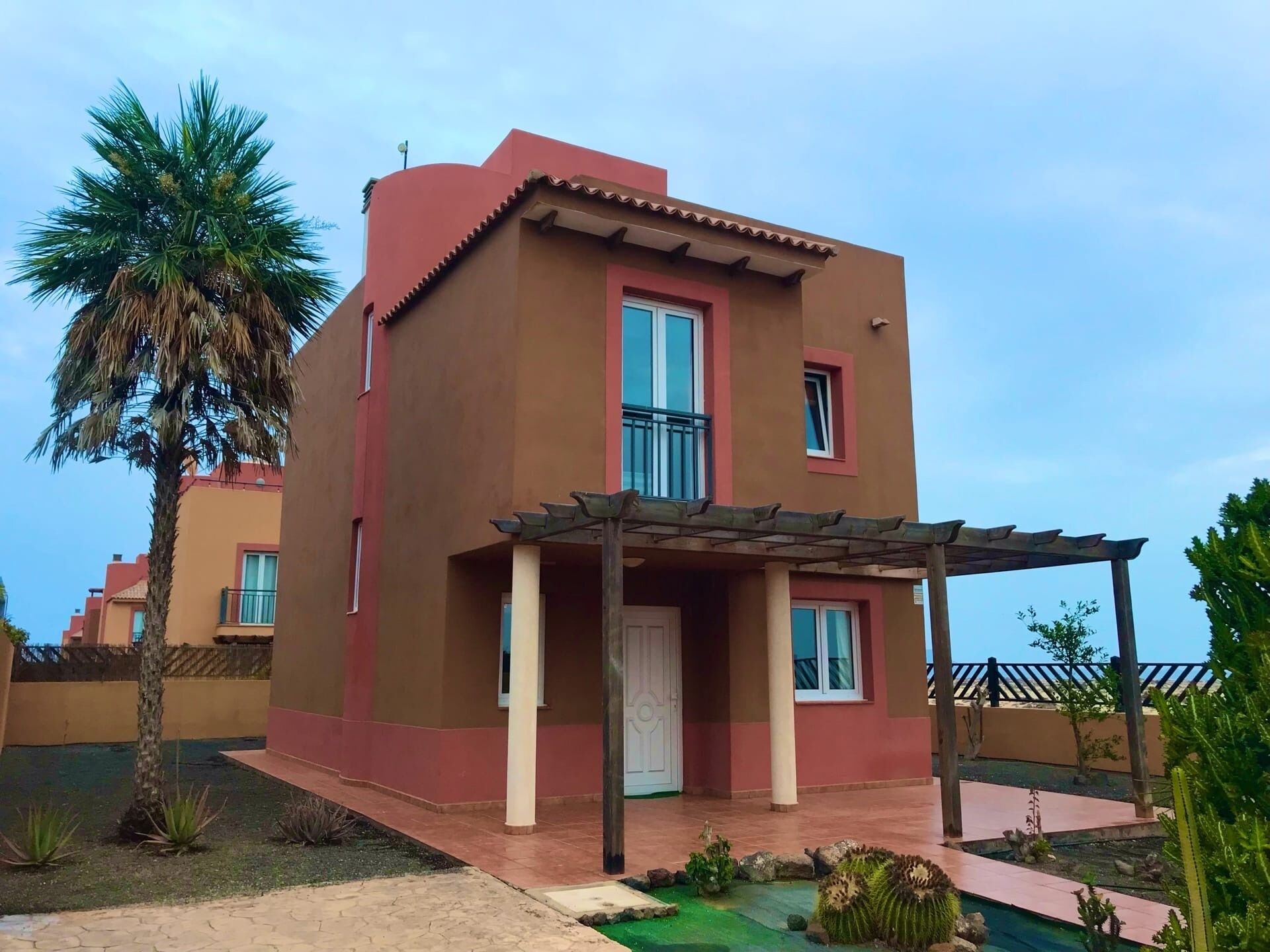 A two-story building with a red and beige facade, a covered entrance, and a palm tree in the foreground.
