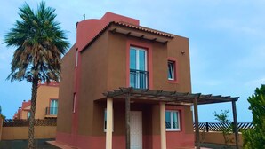 A two-story building with a red and beige facade, a covered entrance, and a palm tree in the foreground. - Relaxing and Welcoming Villa, Corralejo, Canaries (La Oliva)