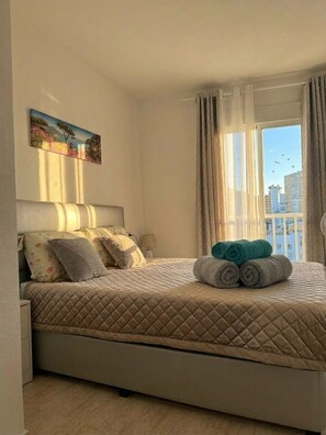 A bedroom with a bed, pillows, a painting, a window with curtains, and a view of buildings. - Godisa 18 Apartment Playa del Cura Torrevieja! (Torrevieja)
