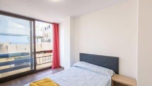 A hotel room with a bed, a wooden nightstand, a large window with a view of buildings and a body of water. - 3 Bed Apartment Sleeps 6 - Free Parking & Balcony (Estepona)