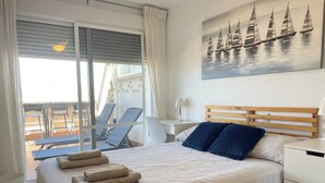 Una camera d'albergo con un letto, un quadro con barche a vela, un balcone con sedie e vista sul mare.