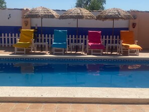 Una piscina con quattro lettini gialli, blu, rosa e arancioni, ognuno con un ombrellone di paglia.