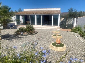 Un edificio con tetto in tegole e ampie finestre, circondato da un giardino ghiaioso con fioriere a forma di fungo e fiori blu.