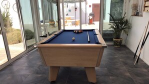 A pool table in a room with dark tiled flooring and large windows.