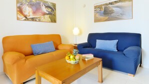 Un salon avec deux canapés, un orange et un bleu, une table basse en bois sur laquelle sont disposés des fruits, et deux tableaux accrochés au mur.