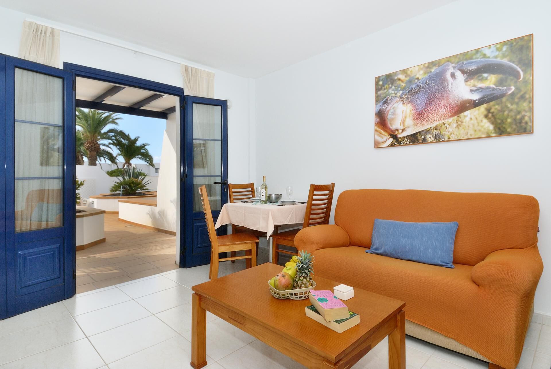 A living room with a sofa, a coffee table with fruit and books, a dining area with a table and chairs, and a large window with curtains.