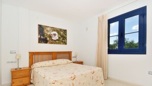 Une chambre à coucher avec un lit, une table de chevet, une lampe, un tableau, une fenêtre avec des rideaux et une vue sur la verdure à l’extérieur.