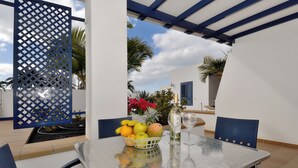 Une table avec un panier de fruits, une bouteille et un verre, placée dans un espace extérieur abrité, offrant une vue sur des palmiers et un ciel dégagé.