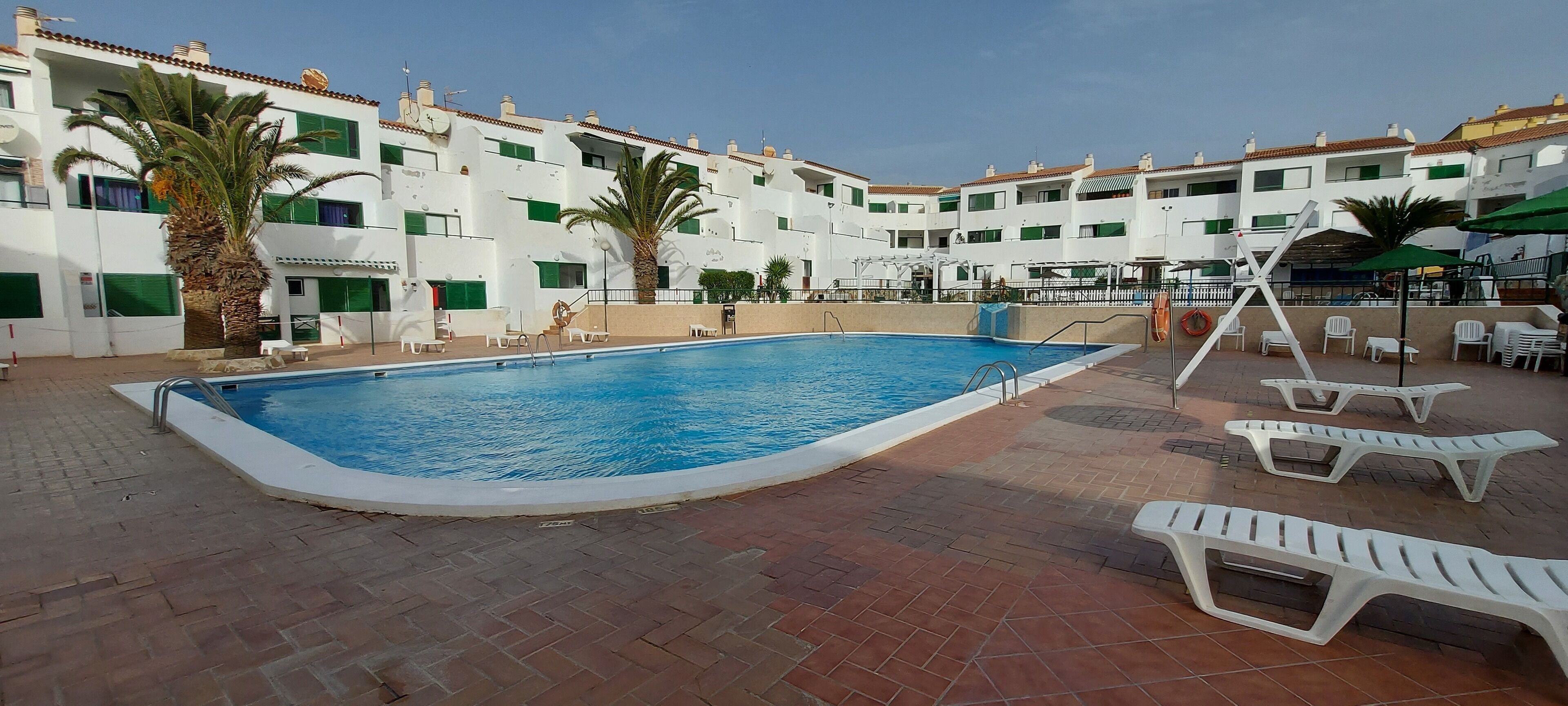 A swimming pool with a tiled deck, surrounded by white buildings with green shutters, palm trees, and outdoor seating.