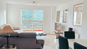 A modern living room with a grey sofa, a dining table with chairs, a TV, and a large window with a view of greenery. - Albatross Golf Suite Alcaidesa (San Roque)