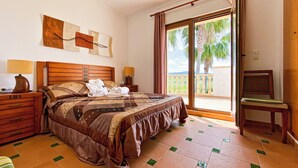 Une chambre d’hôtel avec un grand lit, une tête de lit en bois, deux tables de chevet avec des lampes, une vue sur les palmiers à travers une porte-fenêtre coulissante et un balcon.