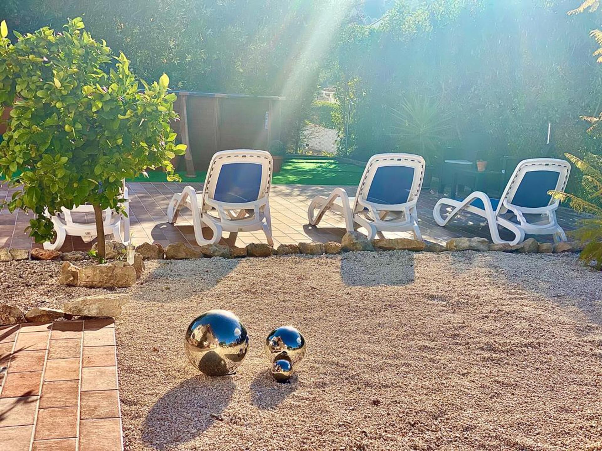A patio area with four white lounge chairs arranged in a row, a small tree, and a decorative spherical object on the ground.
