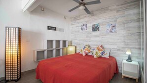 A bedroom with a red bedspread, a ceiling fan, and a wall with framed photos. - Lovely Studio in Costa del Silencio Chayofita (Arona)