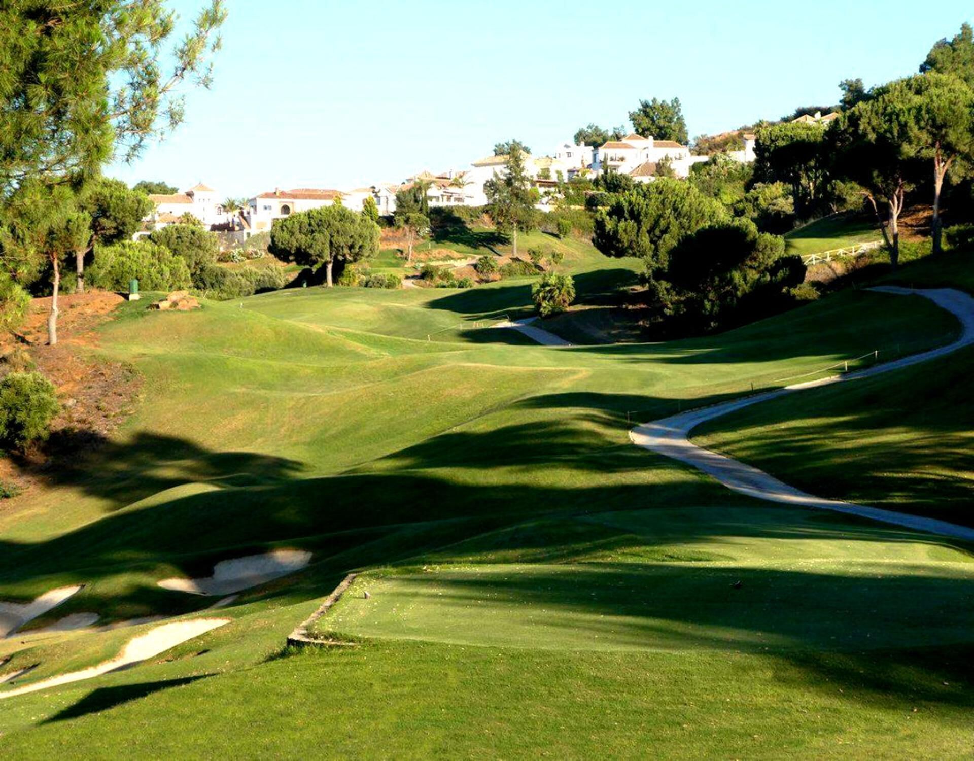 Un campo de golf con un sendero sinuoso, árboles y edificios al fondo.
