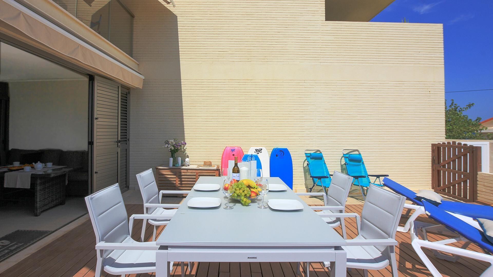 Un patio al aire libre con una mesa dispuesta para comer, rodeado de sillas blancas y sillones azules.